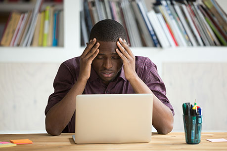 NSA FAQs Part II - a man sits at the desk with a laptop in front of him and his hands on his forehead