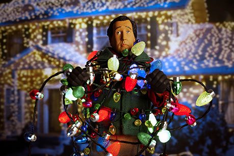 Christmas Vacation character Clark Griswold wrapped in Christmas lights in front of their house 