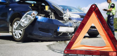 Crashed car with a traffic warning cone in front on the road and a police officer writing a ticket to another car in the background