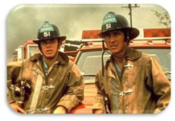 2 firefighters stand in front of a firetruck with smoke in the background