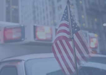 A small, tattered American flag hangs from a pole, with the blurred emergency lights of an ambulance visible in the background.
